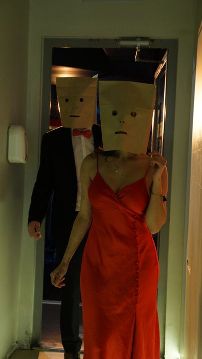 Dos personas con bolsas de papel en la cabeza; una lleva un vestido rojo.